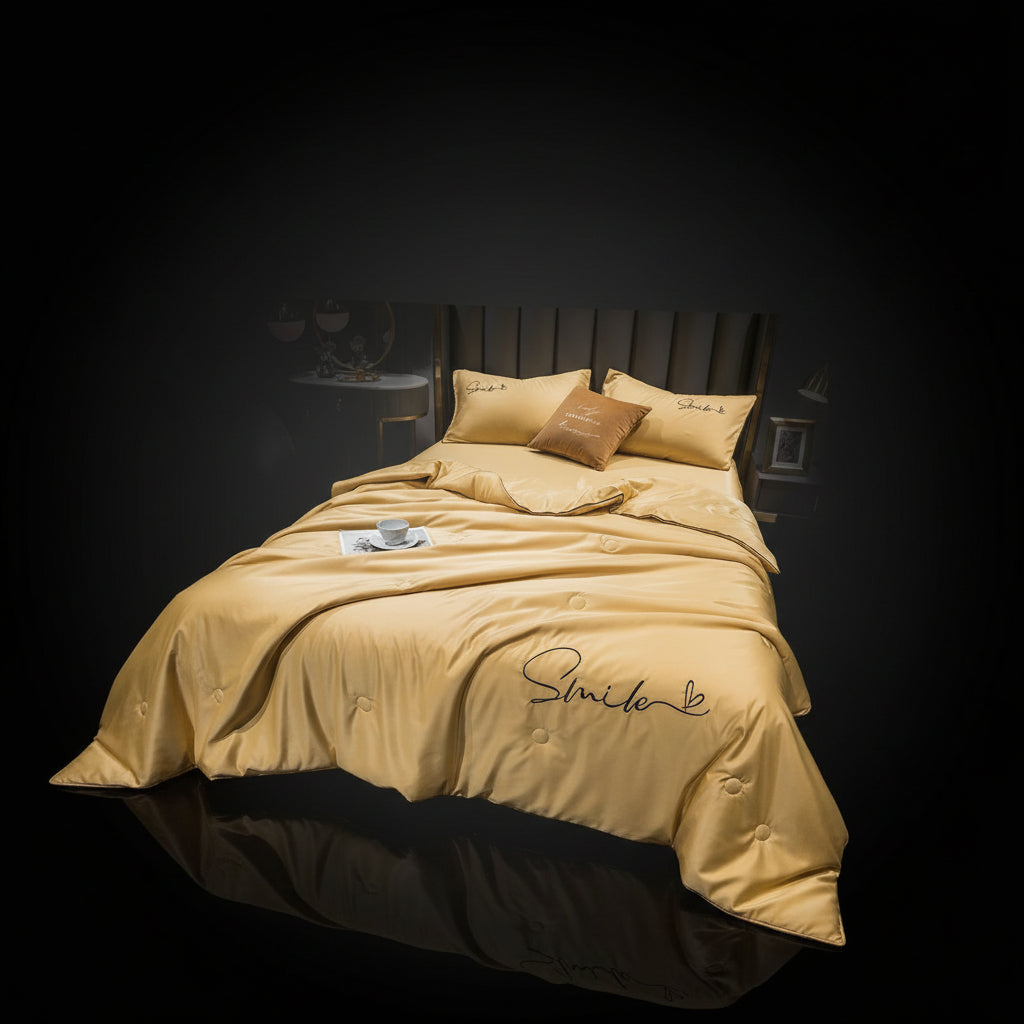 A gray silk summer blanket with embroidery on it, displayed on a bed alongside pillows and a nightstand with a lamp.