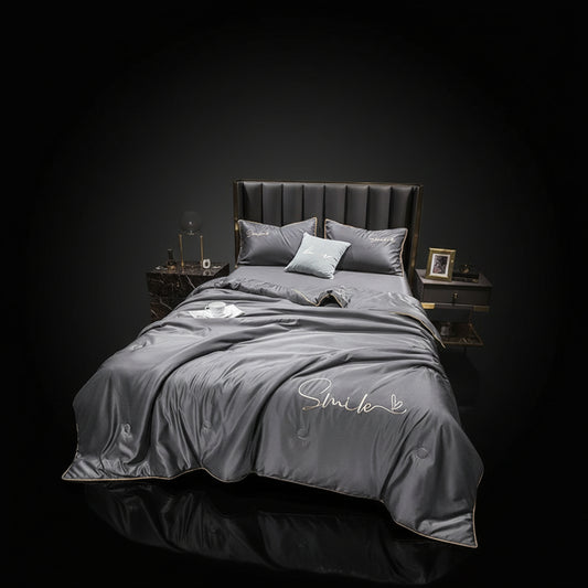 A gray silk summer blanket with embroidery on it, displayed on a bed alongside pillows and a nightstand with a lamp.