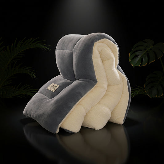 A person sitting on a bed with a dark gray duvet cover, with pillows, a remote control, and a mobile phone on the bed. The room includes a lamp and a plant in the background.