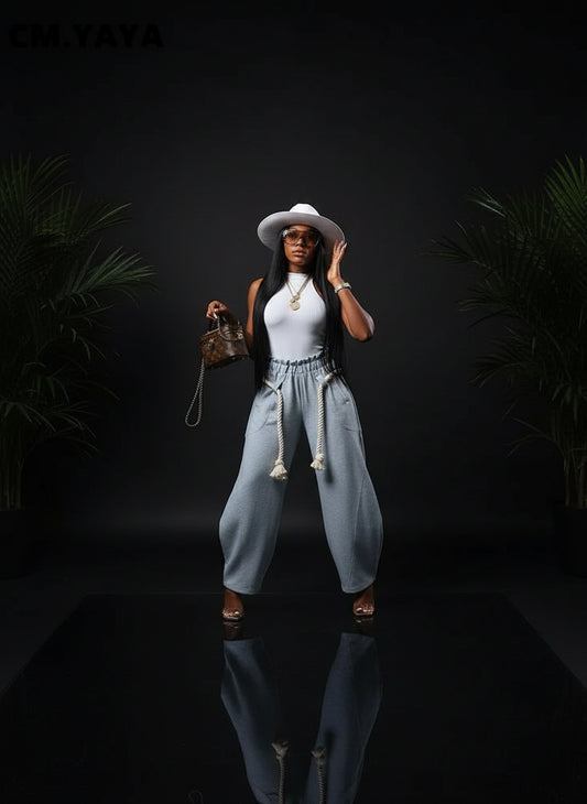 A woman wearing a wide-leg loose jogger pants in gray with pockets and a sash, paired with a white top, hat, and carrying a bag.