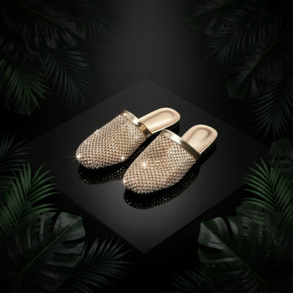 A pair of gold-colored flat slippers adorned with sparkling rhinestones, displayed alongside a pair of blue denim jeans, with the slippers resting on a reflective surface.